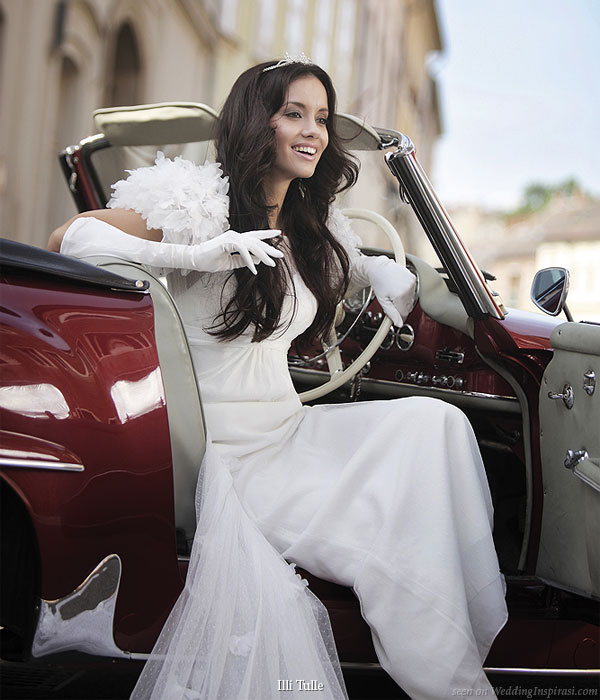Ruffle shoulder bride smiling at the driver's seat of a red vintage car wearing an Illi Tulle Complicite wedding gown